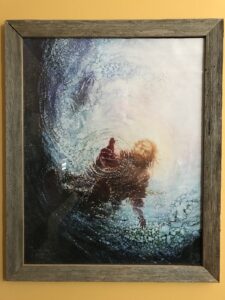 A wall picture with wooden frame of Jesus standing on the water in bare feet reaching into the water to catch Peter